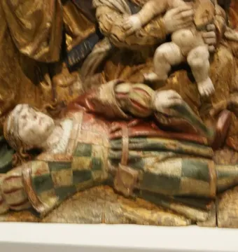 A carved wooden figure from The Last Judgement lies on the ground with arms outstretched, beneath other robed figures on a textured golden background.