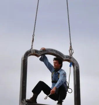 A large dark metal frame for Memoria Urbana hangs from chains while a worker crouches inside it with a wrench.
