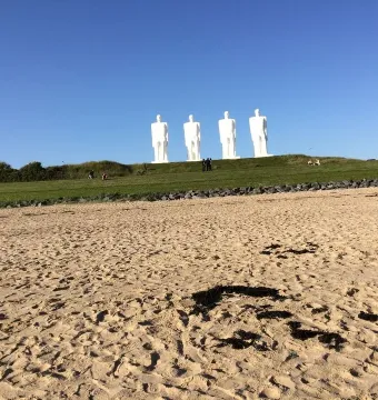 The Man by the Sea sculpture on a grassy hill above a sandy beach, with several visitors nearby.