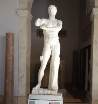 Apoxyomenos, a full-length marble statue of a nude male figure with his arms bent across his chest in a museum gallery.