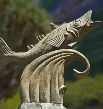 The Chicharro Sculpture shows a metallic fish leaping from a stylized wave outdoors.