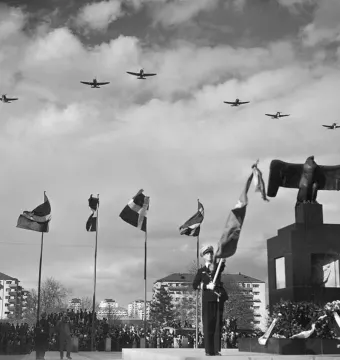Flygarmonumentet with a tall pedestal topped by an eagle sculpture, while eight airplanes fly overhead during a ceremonial gathering.