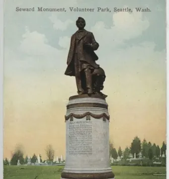 William Henry Seward bronze statue on a stone pedestal in a park, with trees and gravestones in the background.