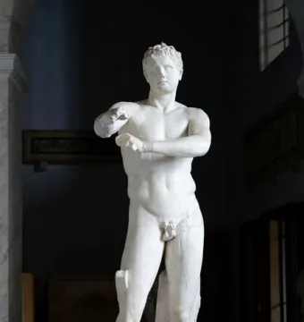 Apoxyomenos marble statue of a nude male figure scraping his forearm in a dim museum hall