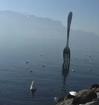 The Fork of Vevey sculpture rises from the water with mountains and birds in the background.
