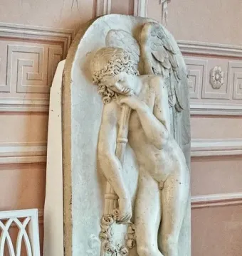 Marble bas-relief of a winged nude male figure leaning on a staff with a floral wreath, set against a pink decorative wall.