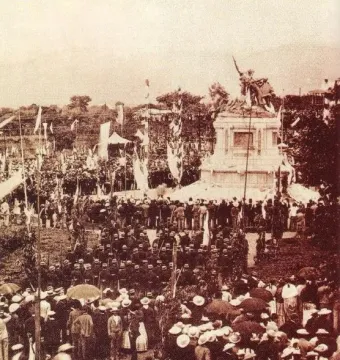The National Monument of Costa Rica stands on a tiered base, surrounded by a large crowd and flags during a public ceremony.