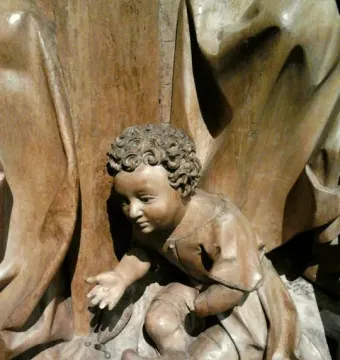 Wooden sculpture of Saint Luke as a curly-haired child kneeling with one hand extended.