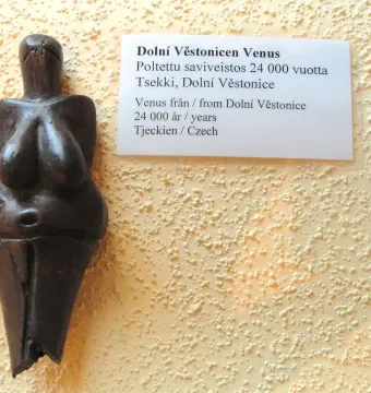 Venus of Dolní Věstonice figurine with prominent breasts and a rounded belly, displayed beside a multilingual label.