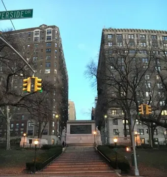 The Firemen\'s Memorial beside a broad stone stairway between two apartment buildings, with bare trees and a crosswalk in front.
