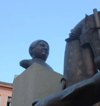 Dark metallic statue of Jaroslav Hašek with a horse head in the foreground and a bust on a stone pedestal against a blue sky.