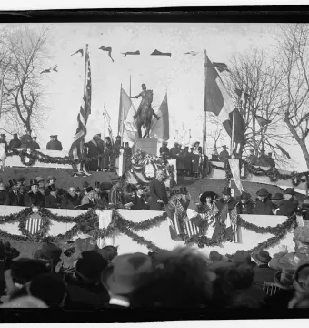 Joan of Arc equestrian statue on a pedestal at an outdoor gathering, with flags, garlands, spectators, and bare trees around it.