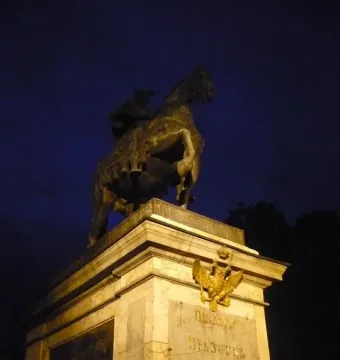 Illuminated bronze equestrian monument to Peter I on a tall stone pedestal with a double-headed eagle and Cyrillic inscriptions.