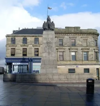 Paisley War Memorial stone cenotaph in a town square with carved details and dark figures on top.