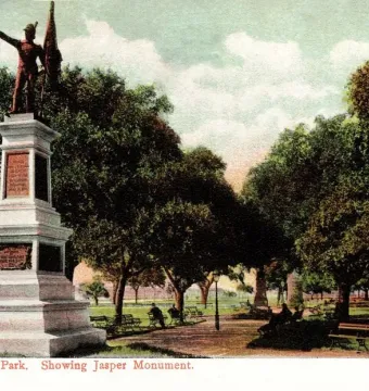 The Defenders of Fort Moultrie monument rises on a stone pedestal in a park with trees, paths, and benches.