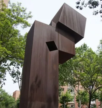 El Llarg Viatge, a tall abstract reddish-brown metal sculpture with a square opening, standing in an urban park.