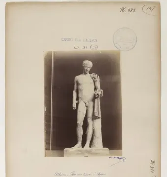 White marble statue of Hermes of Aegium in a nude contrapposto pose with drapery over one shoulder and a support at his side.