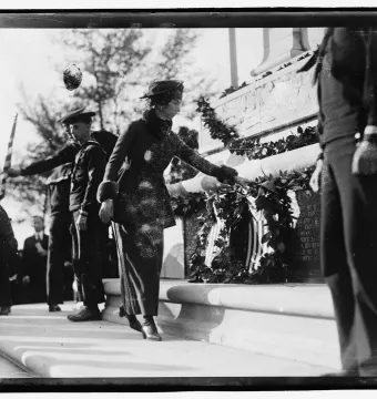 A woman places a wreath on the John Paul Jones Memorial while men in naval uniforms stand nearby on the steps.