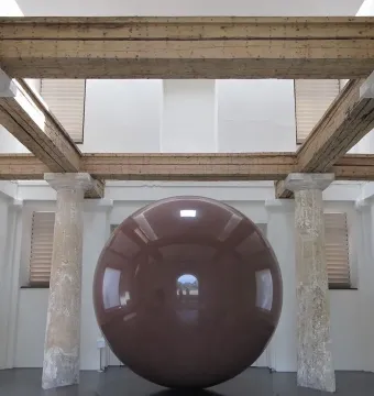Large Red Sphere displayed indoors between weathered classical pillars.