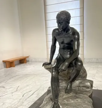 Seated Hermes, a dark bronze nude male statue on a black plinth in a museum room.