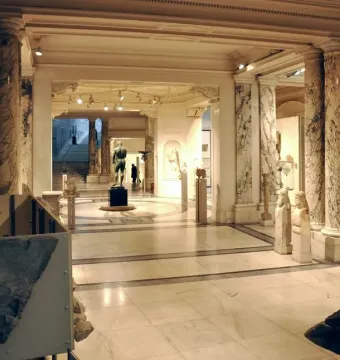 The Athlete from Ephesus standing on a pedestal in a columned museum hall, seen from behind.