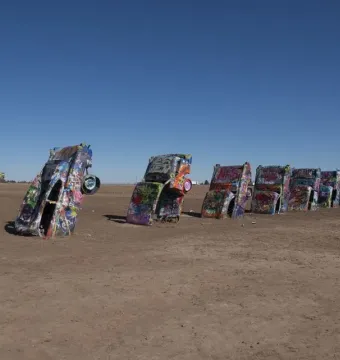 Cadillac Ranch cars covered in graffiti stand buried nose-first in a flat dirt field under a clear blue sky.