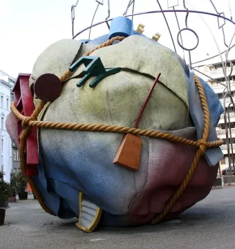 Houseball sculpture with a large textured sphere in yellow, blue, and red, wrapped with a rope and attached abstract forms.