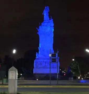 The Monument to the Carta Magna and Four Regions of Argentina lit in vivid blue at night, with a small stone pillar, streetlights, and trees nearby.