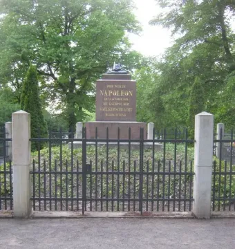 Napoleonstein stone monument behind a dark metal fence and stone pillars, surrounded by trees and greenery.