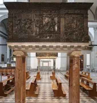 The Passion Pulpit in a church interior, with dark bronze relief carvings on red marble columns above a checkered floor and wooden pews.