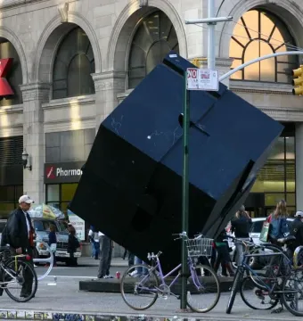 The Alamo sculpture, a large dark tilted cube, stands in an urban street scene near a stone building and street signs.