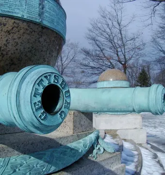 Close-up of two bronze cannons at the Battle Monument, with GETTYSBURG visible on the front cannon and a snow-dusted monument behind them.