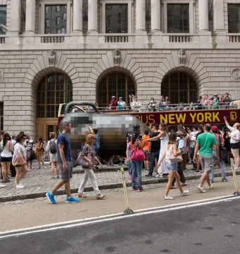 Charging Bull sculpture on a city street surrounded by pedestrians, with a tour bus and stone building in the background.