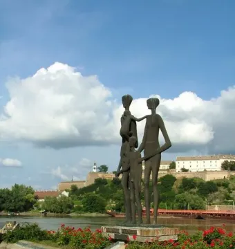 Monument to the Victims of Fascism with bronze figures on a brick platform beside flowering bushes and a river.