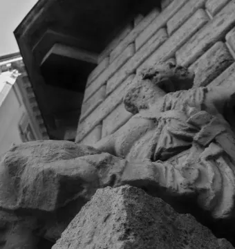Weathered Pasquino stone statue with eroded draped figures beside a stone building and a metal rod protruding from the shoulder.