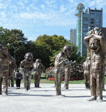 A-maze-ing Laughter sculpture with metallic human-like figures in an outdoor plaza, with buildings and trees behind it.