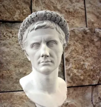 Bust of Augustus wearing a gemmed crown, displayed on a blue stand against a stone wall.