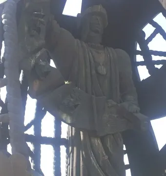 Detail of the Birth of a New Man sculpture showing a dark figure holding a scroll, framed by a rope lattice and ship mast.