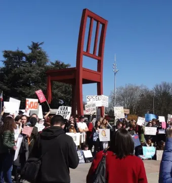 The Broken Chair sculpture stands tall in red wood behind a crowd of protesters holding signs.