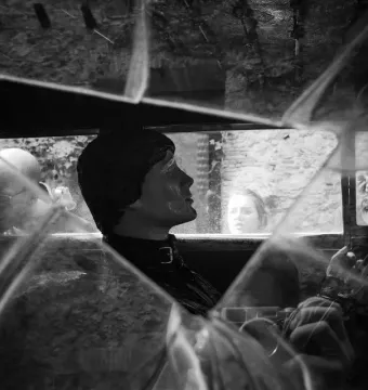 Surreal black and white scene of Rainy Taxi seen through a fractured windshield, with a mannequin in the driver\'s seat and reflections of two people in the windows.
