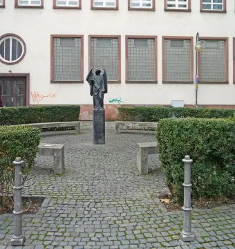 The Frankfurter Engel statue stands on a pedestal in a paved plaza with hedges and benches in front of a building with a circular window.