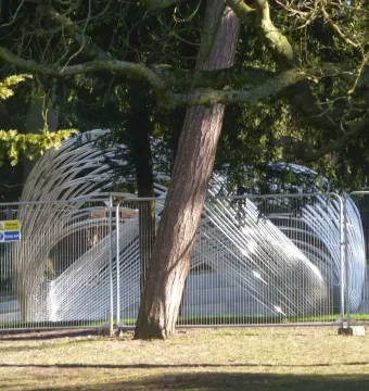 Infinite Wave sculpture behind a temporary metal fence in a wooded park setting.