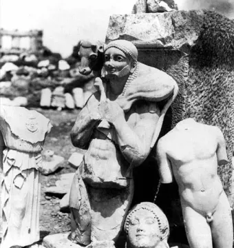 A headless nude marble torso, identified as the Kritios Boy, standing among fragmented ancient statues and ruins.