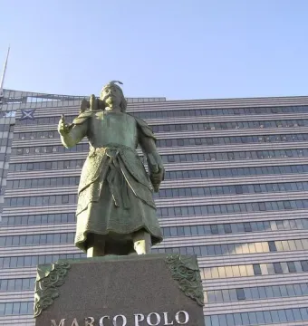 Marco Polo statue in historical attire with a bird on his arm, standing on a stone pedestal.