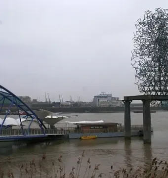 Quantum Cloud sculpture rising above the River Thames near a blue arched bridge and waterfront buildings.