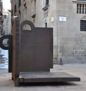 Topos V, a dark geometric sculpture by Eduardo Chillida, stands in a stone-paved courtyard beside old stone buildings.