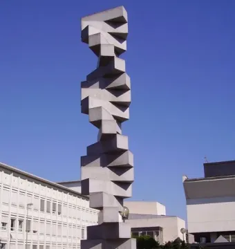 Endlose Treppe sculpture rising on a cylindrical base beside modern buildings under a clear blue sky.