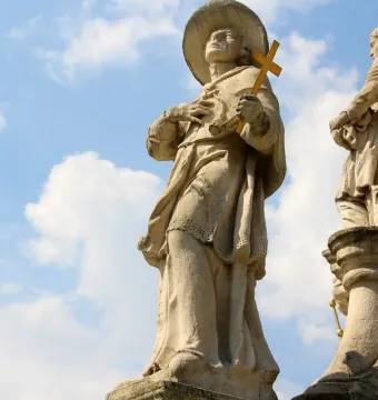 Weathered stone figure from the Holy Trinity Monument holding a golden cross and looking upward, with another robed statue partly visible beside it.
