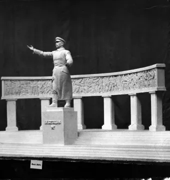 A model for a Stalin monument showing a figure in a long coat and cap before a curved colonnade with relief sculptures.