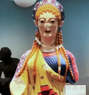 Korè 675, a brightly painted ancient Greek female statue with braided hair and a decorated headdress.
